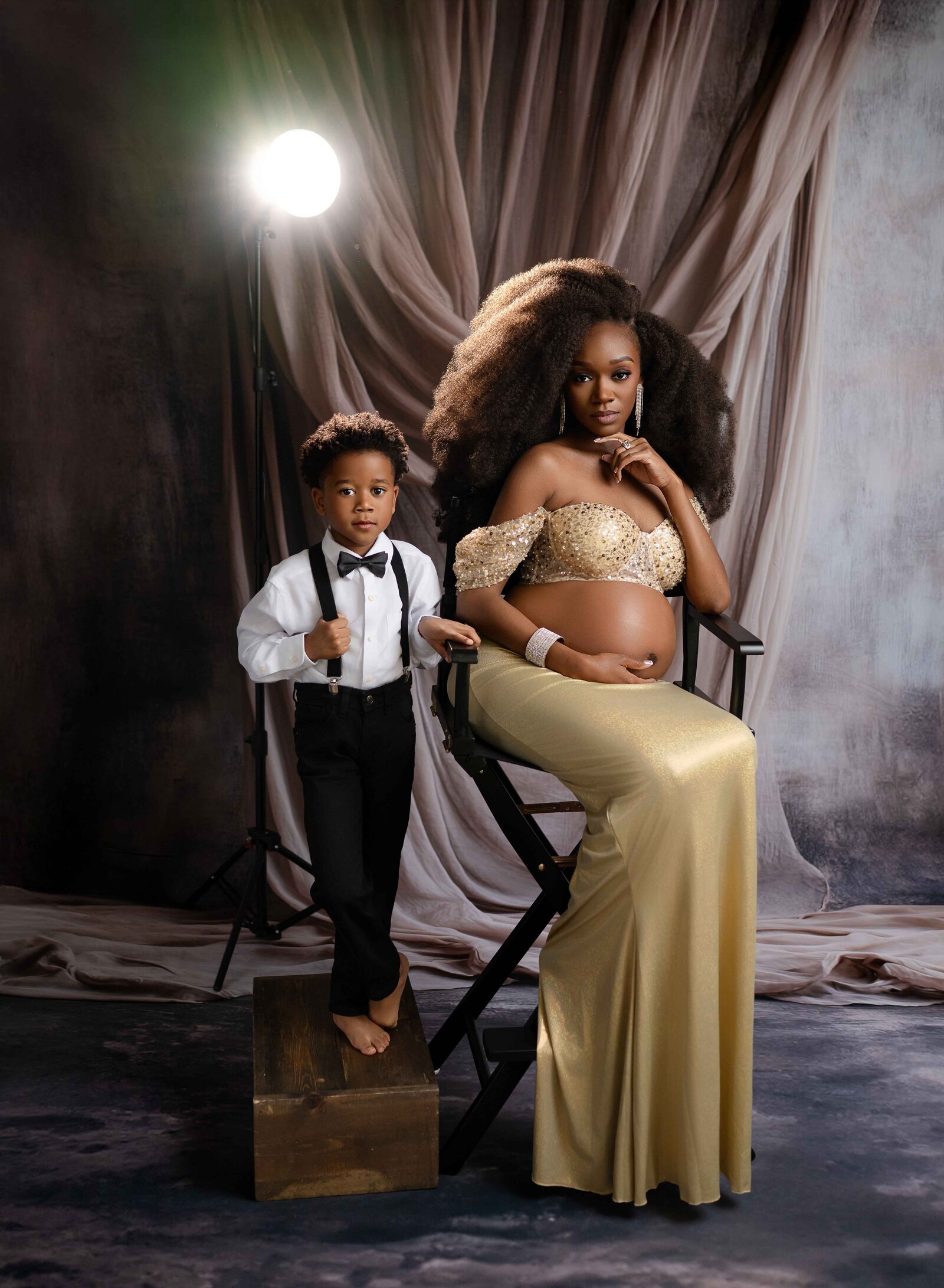Pregnant women in gold gown, sitting in director's chair with son in black suspenders and bow tie. Both looking at camera with a serious look and spotlight behind them. Photographed by Atlanta Maternity photographer.