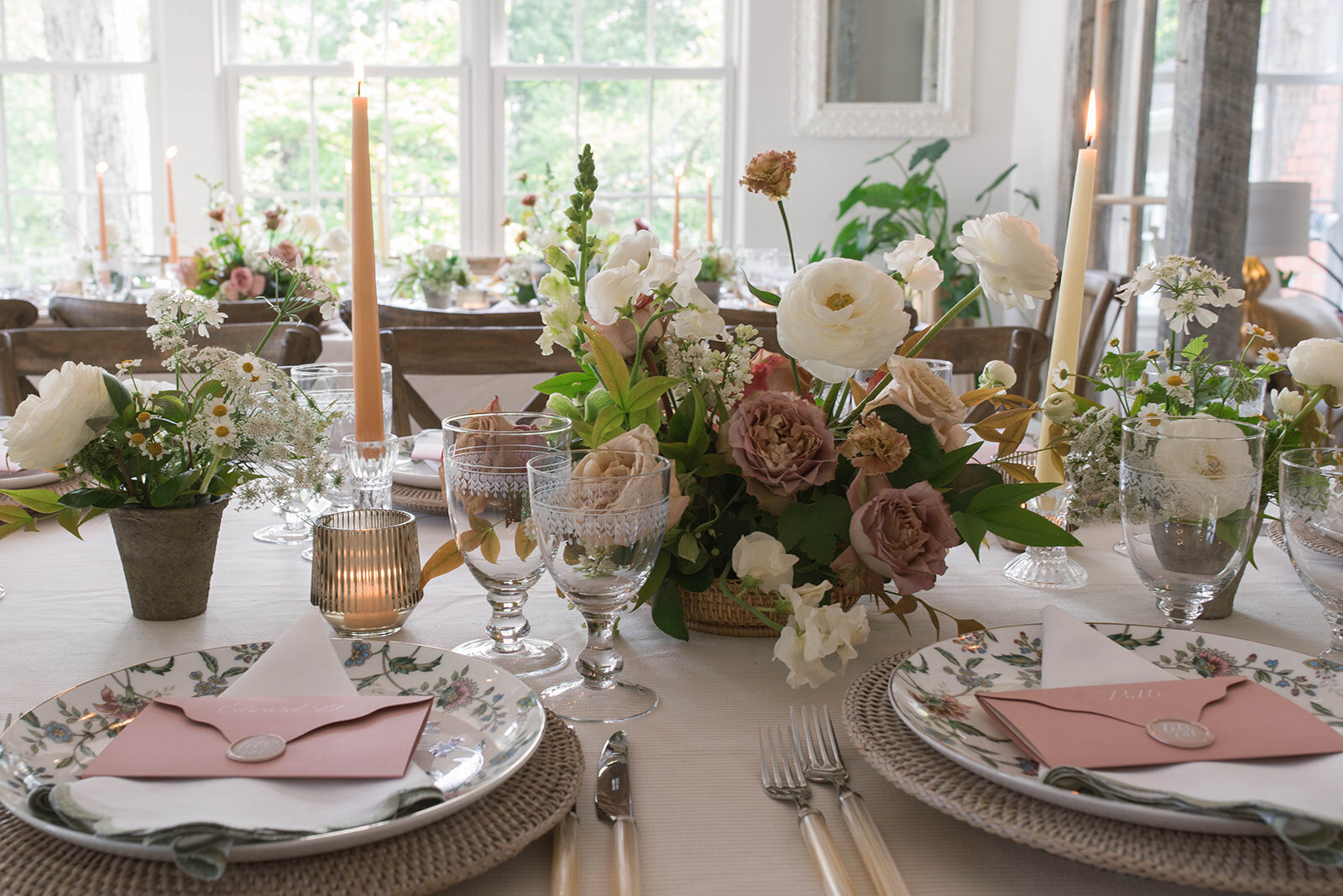 A long table with floral arrangements including white ranunculus, white snapdragons, brown lisianthus, mauve garden roses, and greenery with cream and sand-stone colored taper candles.