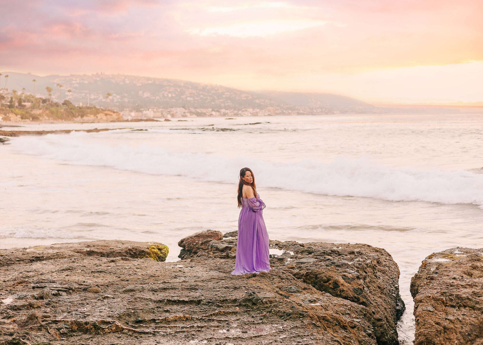 Momma posed in Laguna Beach wearing purple dress during sunset maternity session by Ashley Nicole photography.