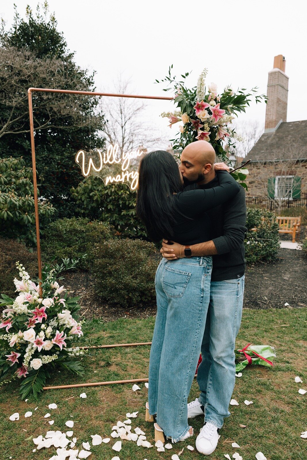 Couple embracing in front of a floral proposal backdrop with a neon 'Will you marry me?' sign, styled by Yours Truly, a wedding planner and designer in New Jersey.