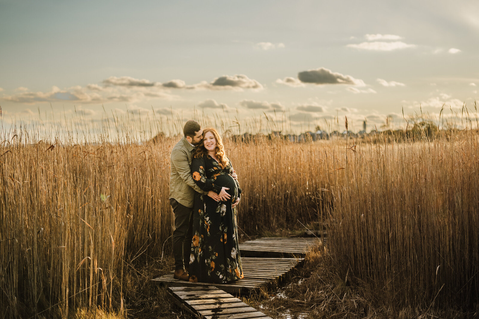 pregnant couple snuggles on a boardwalk at sunset