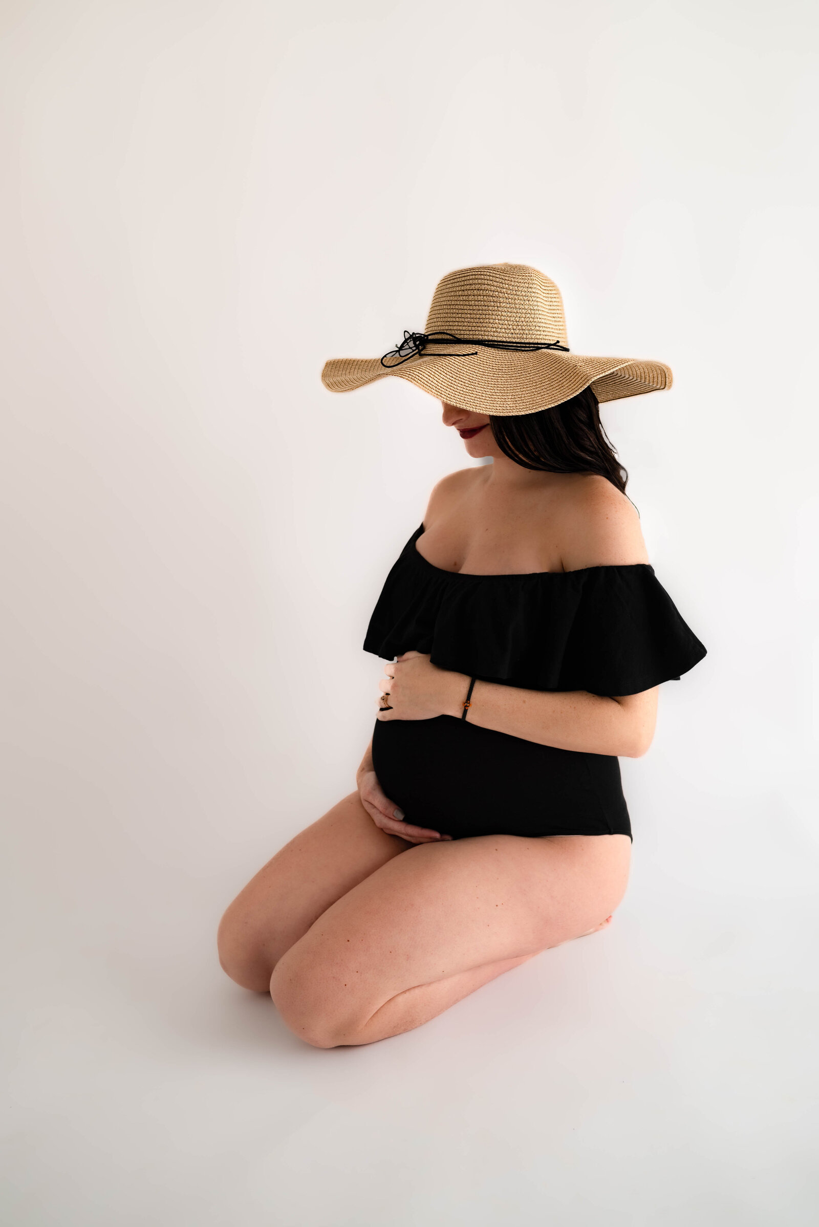 A pregnant woman wearing a black body suit and tan hat kneeling on the floor for her Huntsville Alabama studio maternity pictures