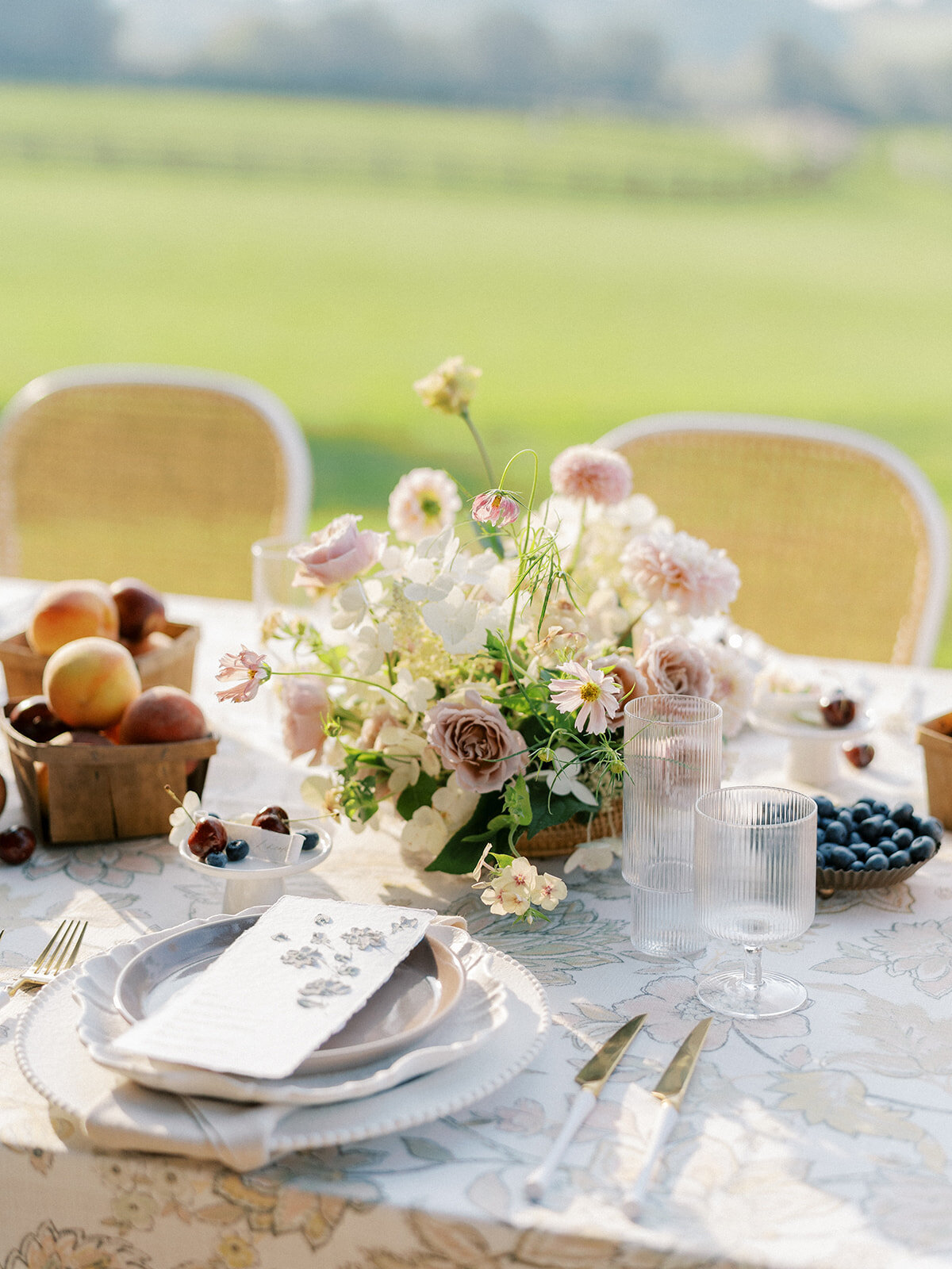 Table setting on top of a floral-patterned linen, with fresh fruit and wispy floral compotes going down the length of the table including taupe and blush garden roses, white hydrangea, brown lisianthus, phlox and blush cosmos.
