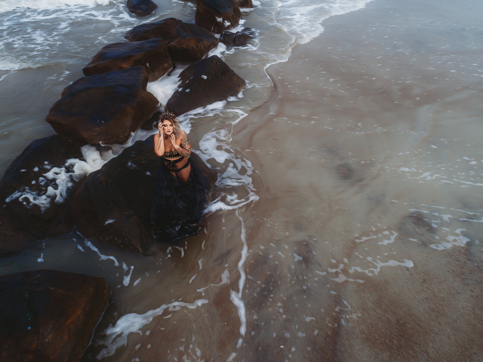 Aerial shot of woman on beach with black gown by rocks hands by face