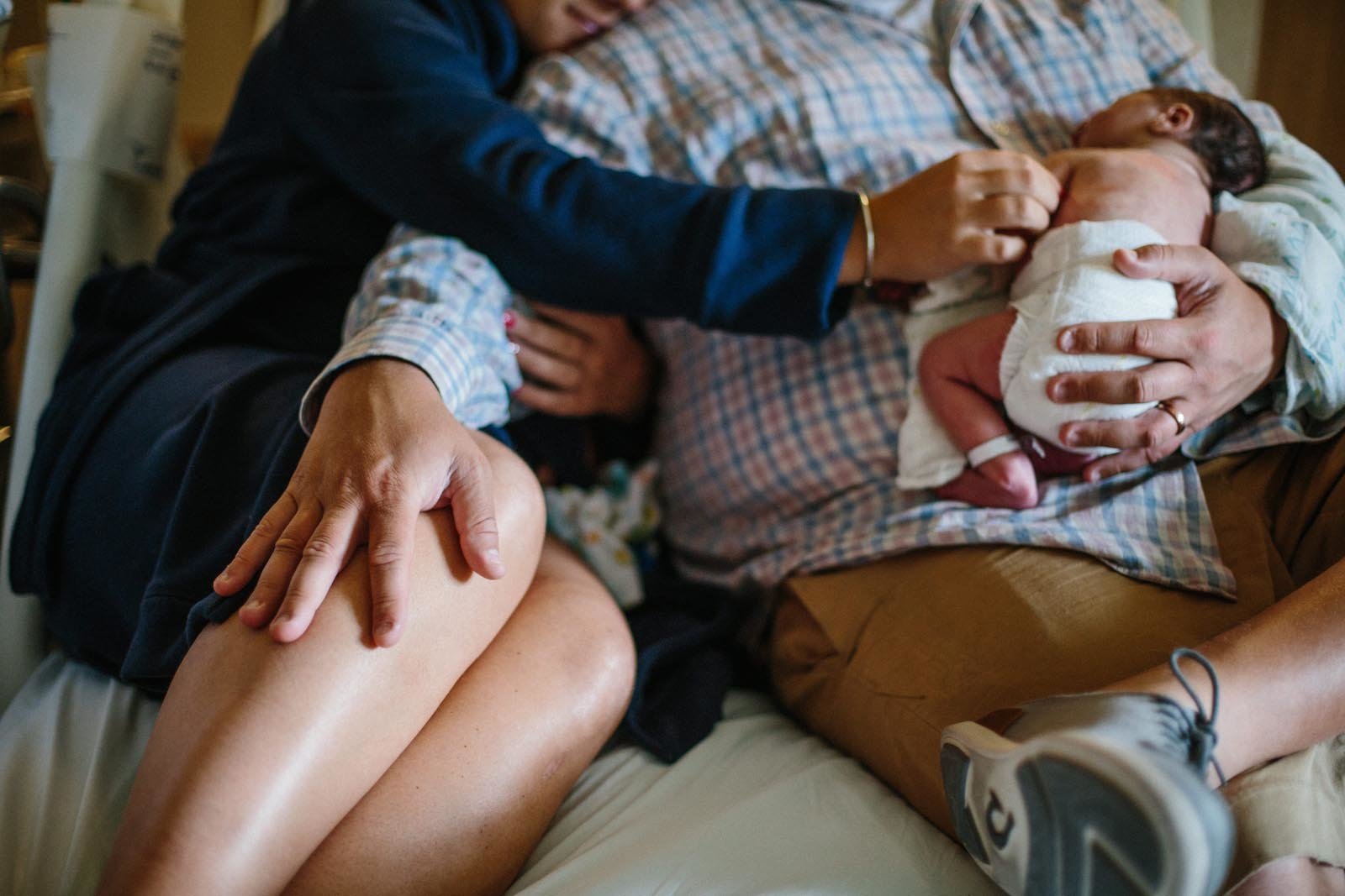 closeup of mom and dad sitting on hospital bed with dad holding newborn baby boy while mom reaches over to place her hand on him