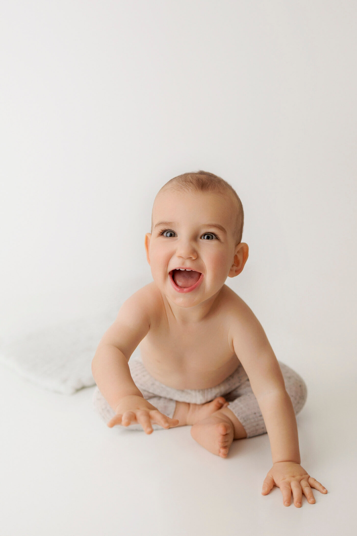 A joyful baby with light hair and big blue eyes sits on a white surface, wearing beige knit pants. The baby is smiling widely with their mouth open, showing excitement and happiness. A soft white blanket is visible in the background, adding to the clean and minimalistic setting.