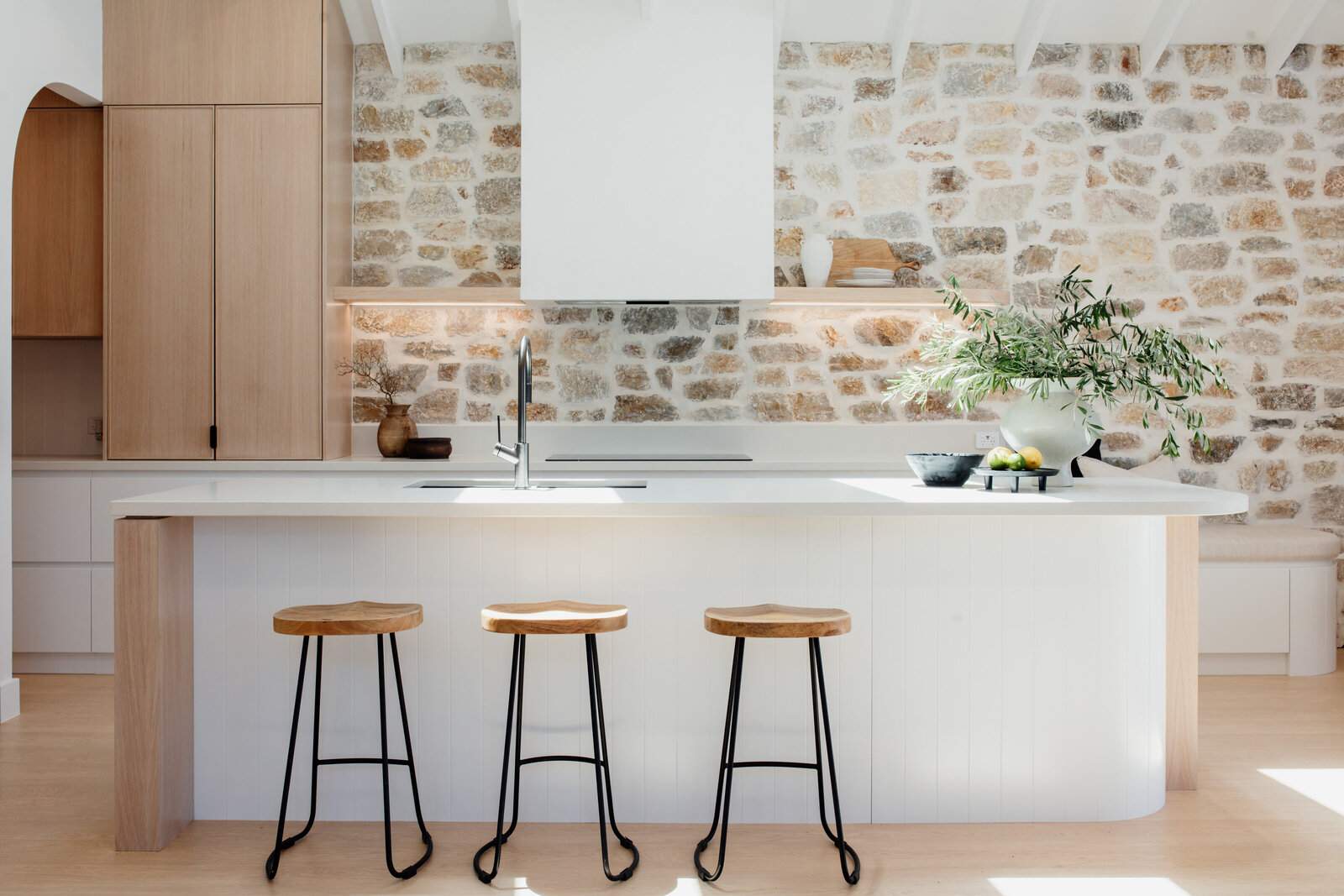 Bright kitchen with stone wall, wooden cabinets, white island, three stools, and green plant decoration. Designed by a Newcastle interior designer specializing in custom projects that transform spaces into stunning showcases of style.