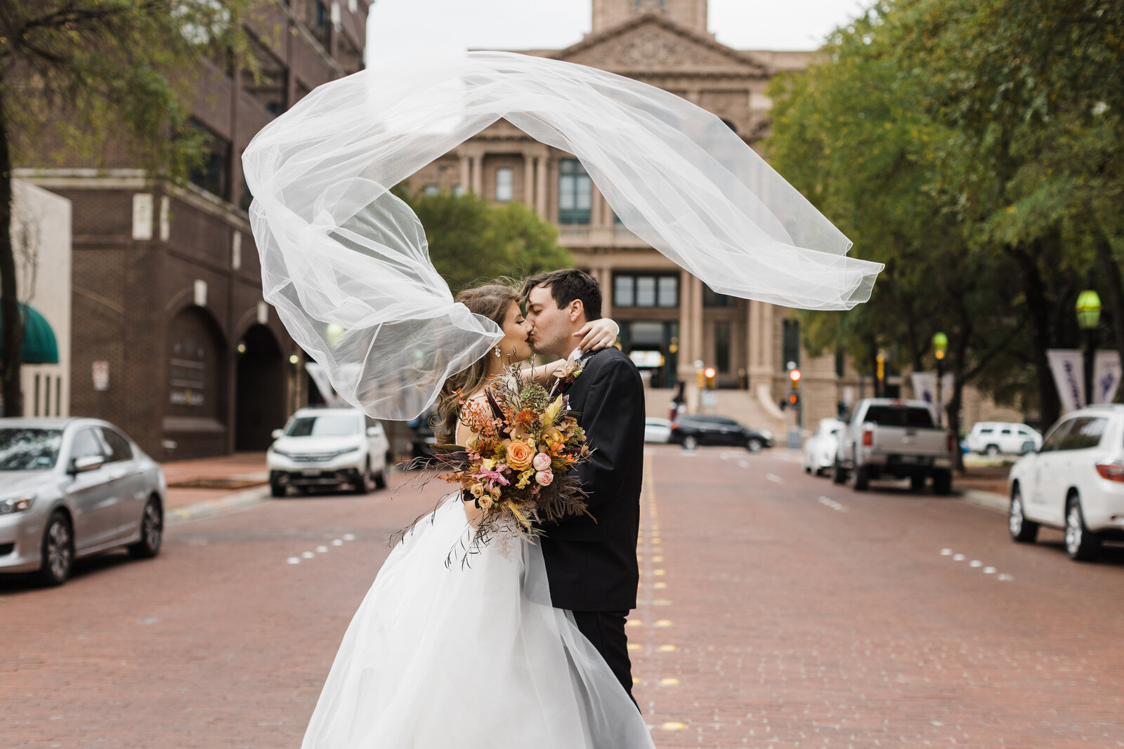 A bride and groom kiss in front of the Tarrant County Courthouse in downtown Fort Worth, Texas. The bride's veil wraps around the couple and perfectly frames their faces in a circular motion. The bride is holding an orange, earthy bouquet and is wearing a full, white dress. The groom is in a black suit. It's a cloudy and windy day.