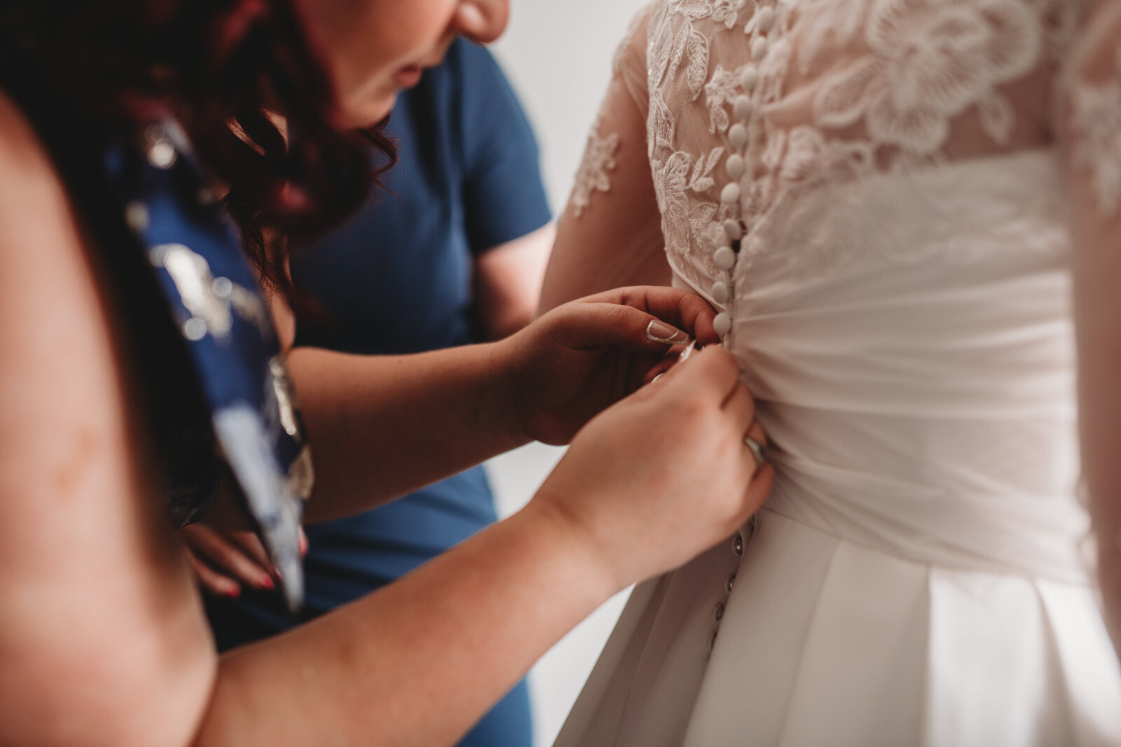 A heartfelt wedding morning moment as a bridesmaid carefully fastens the buttons on a bride's lace gown. This intimate bridal preparation shot captures the beauty and anticipation of a Connecticut wedding day, emphasizing the elegant details and special bonds shared before the ceremony.