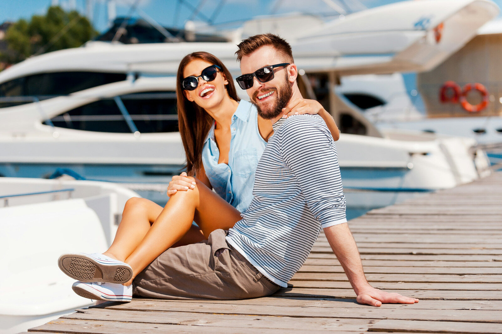 A smiling couple sits on a dock near boats, with the woman wrapping her arm around the man, both are wearing sunglasses and casual clothes