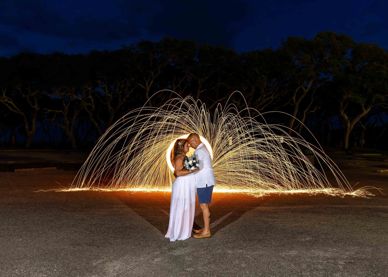 The bride and groom embrace under a dazzling sparkler art display with a backdrop of trees, captured during a Fort Fisher, NC wedding, showcasing magical and creative nighttime photography.