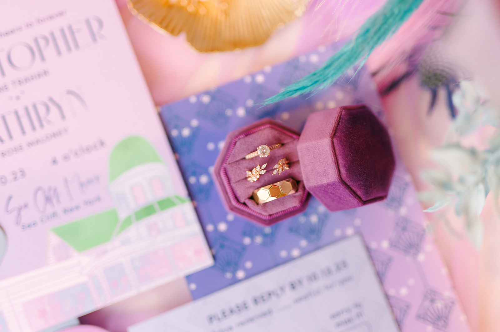 A close up of a gold engagement ring and gold wedding rings in a purple wedding ring box on top of a wedding invitation.