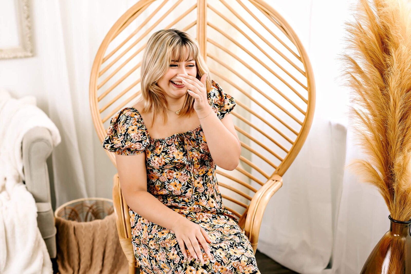 A woman with blonde hair sits in a wicker chair, wearing a floral dress and laughing with her hand over her mouth. The setting is bright and decorated with a white couch and tall, tan pampas grass.