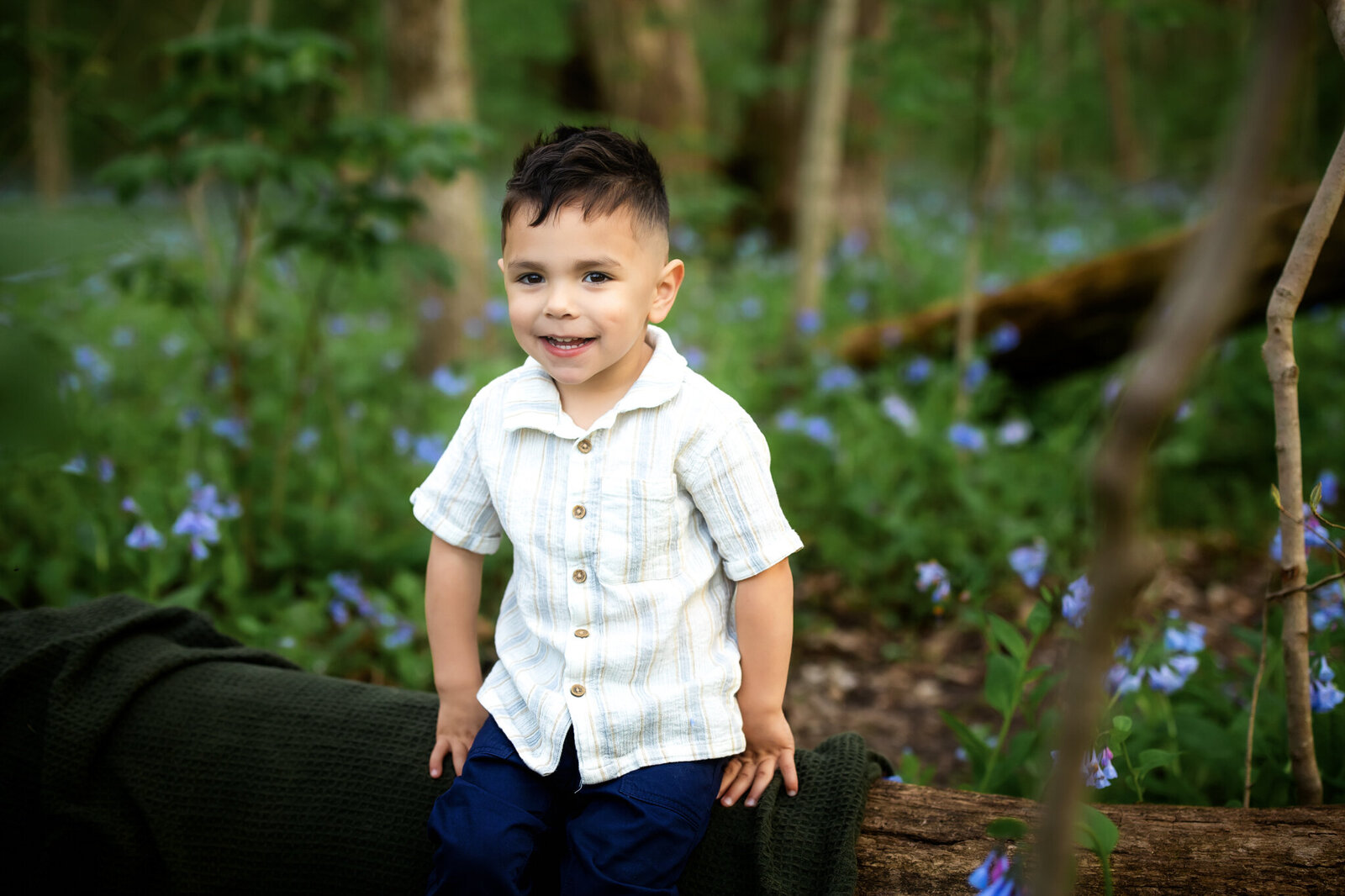 A young boy with short dark hair, wearing a light-colored short-sleeve shirt and navy pants, sits on a log in a lush forest with green foliage and purple wildflowers—captured in stunning outdoor portraits by a Bloomington family photographer.