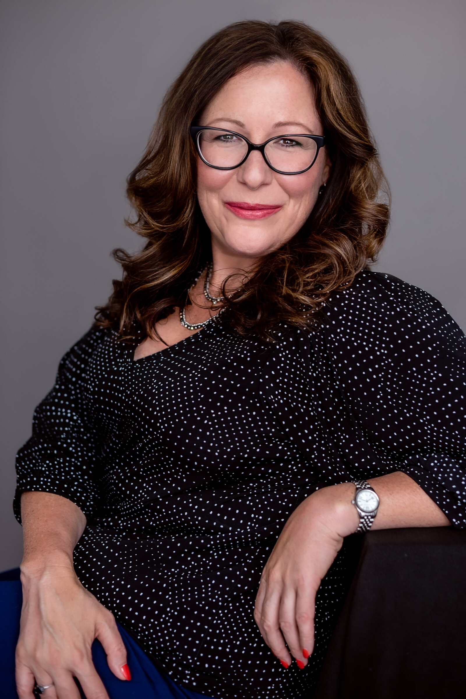 Headshot of an older woman with auburn shoulder length hair poses casually wearing a dark blouse against a grey backdrop