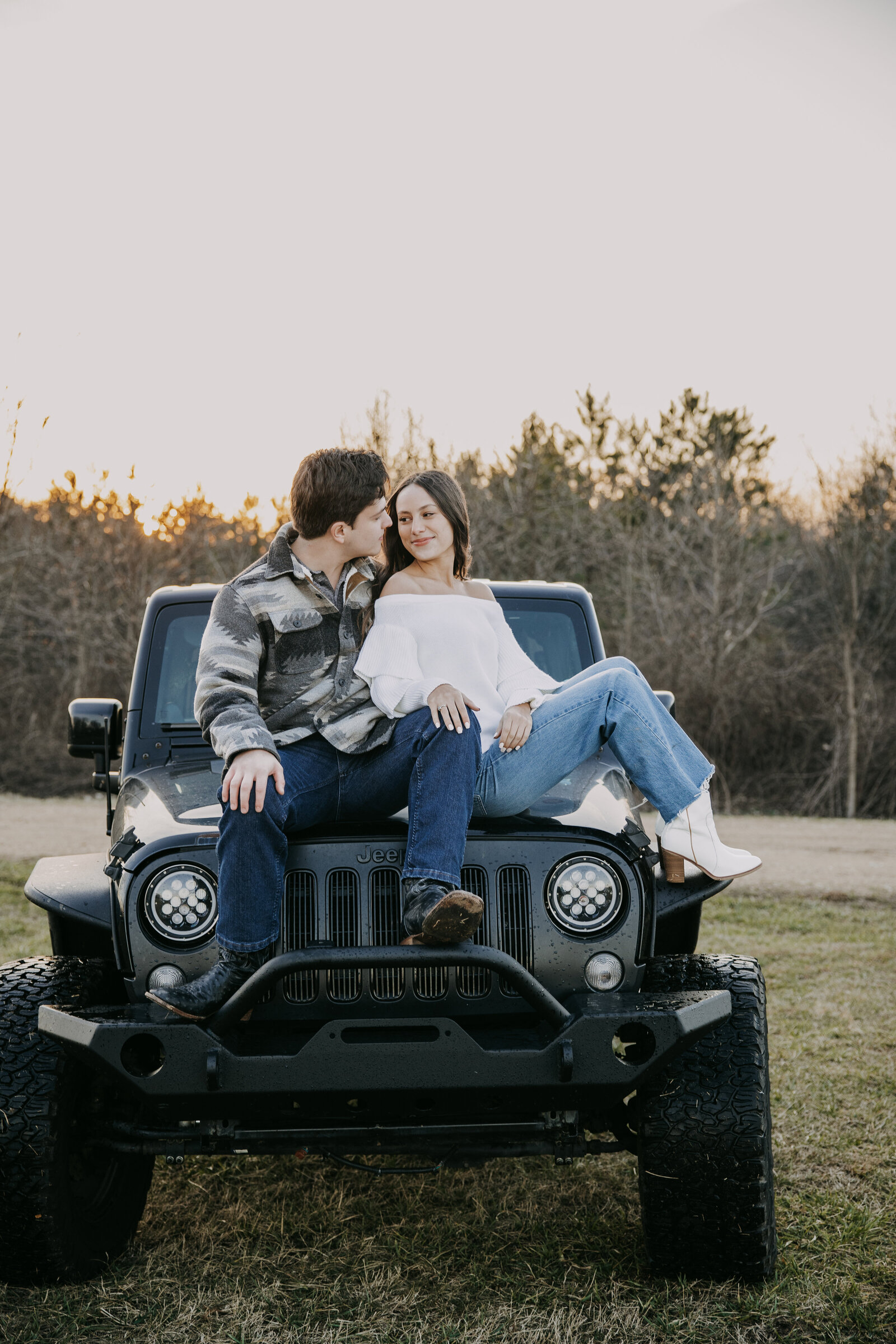 A couple sits on the Jeep’s hood, cuddling closely as the man kisses her lovingly, wrapped in the golden glow of sunset.