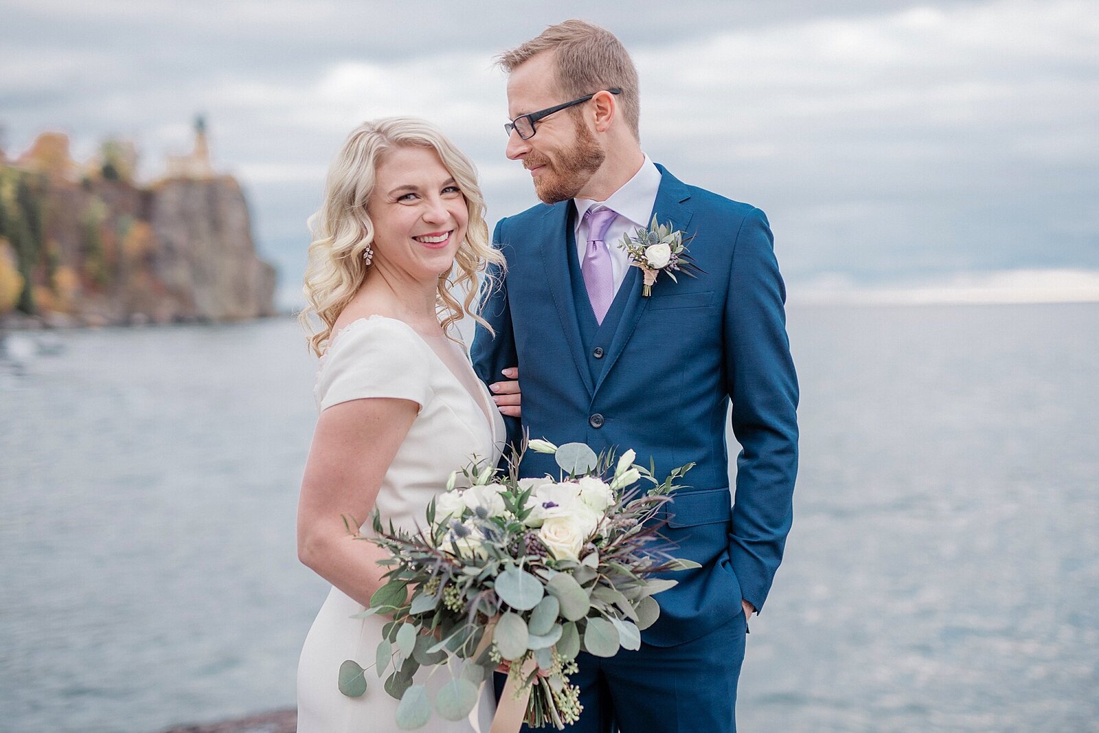 A man in a blue suit smiles down at his wife after their Minnesota wedding along Lake Superior. Split Rock Light House is in the background.