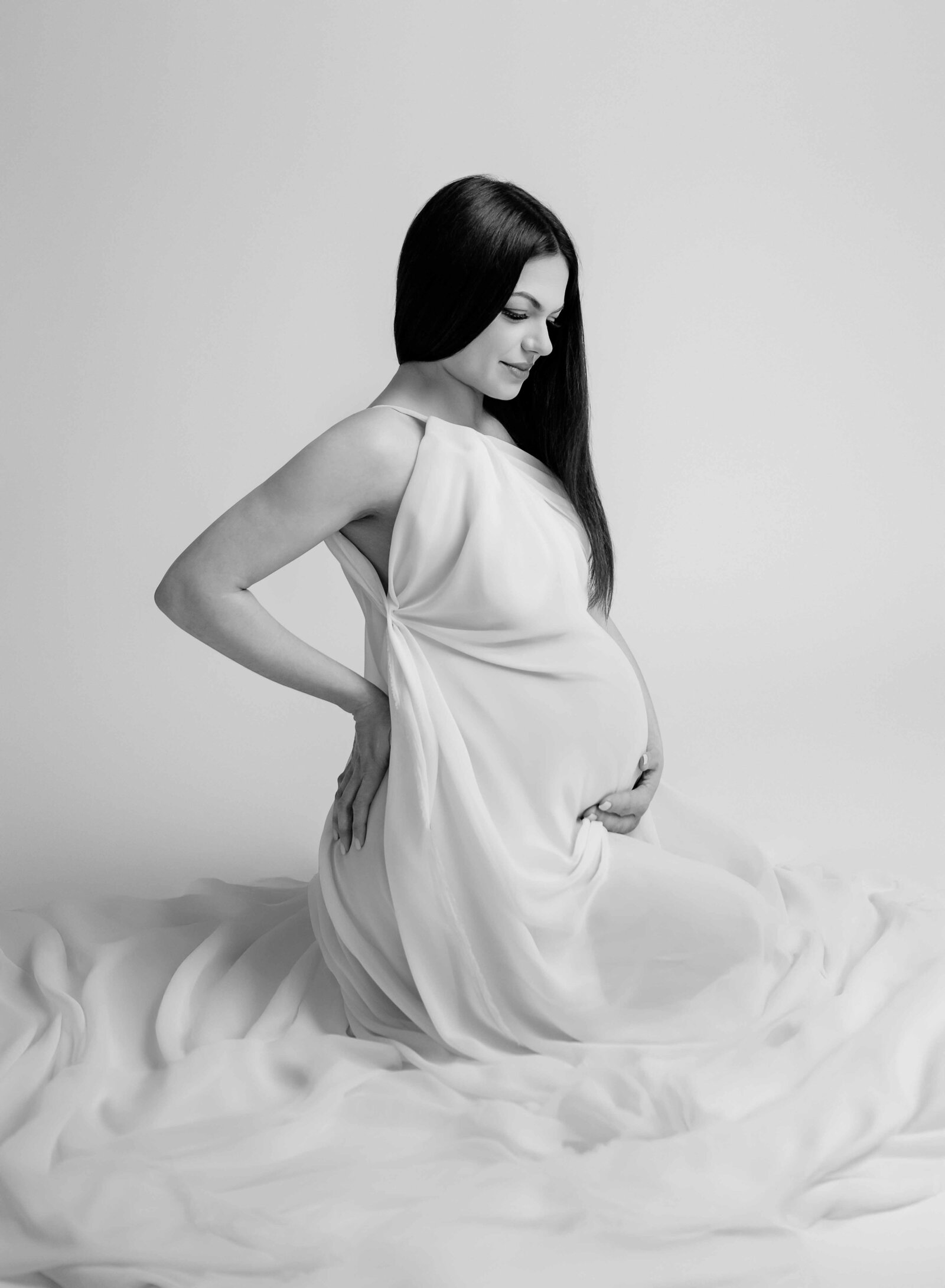 Mom kneeling on ground, wrapped in white drape, looking at belly. Photo taken by Atlanta Maternity Photographer- Jenni Nikki in Woodstock, GA.
