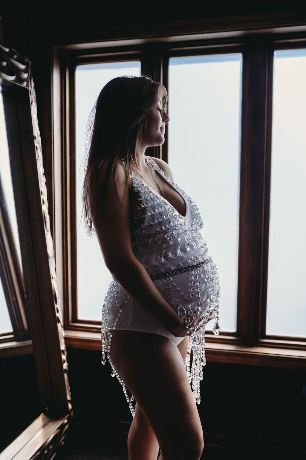 maternity boudoir of woman in white jewel lingerie in front of windows with hand on bump