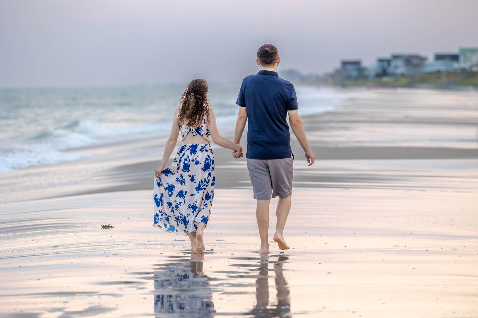 An engaged couple walking hand-in-hand along the shoreline in Wilmington, NC, with waves gently lapping at their feet. This image embodies candid wedding photography, showcasing a beautiful connection that resonates with couples looking for unique wedding photography packages.