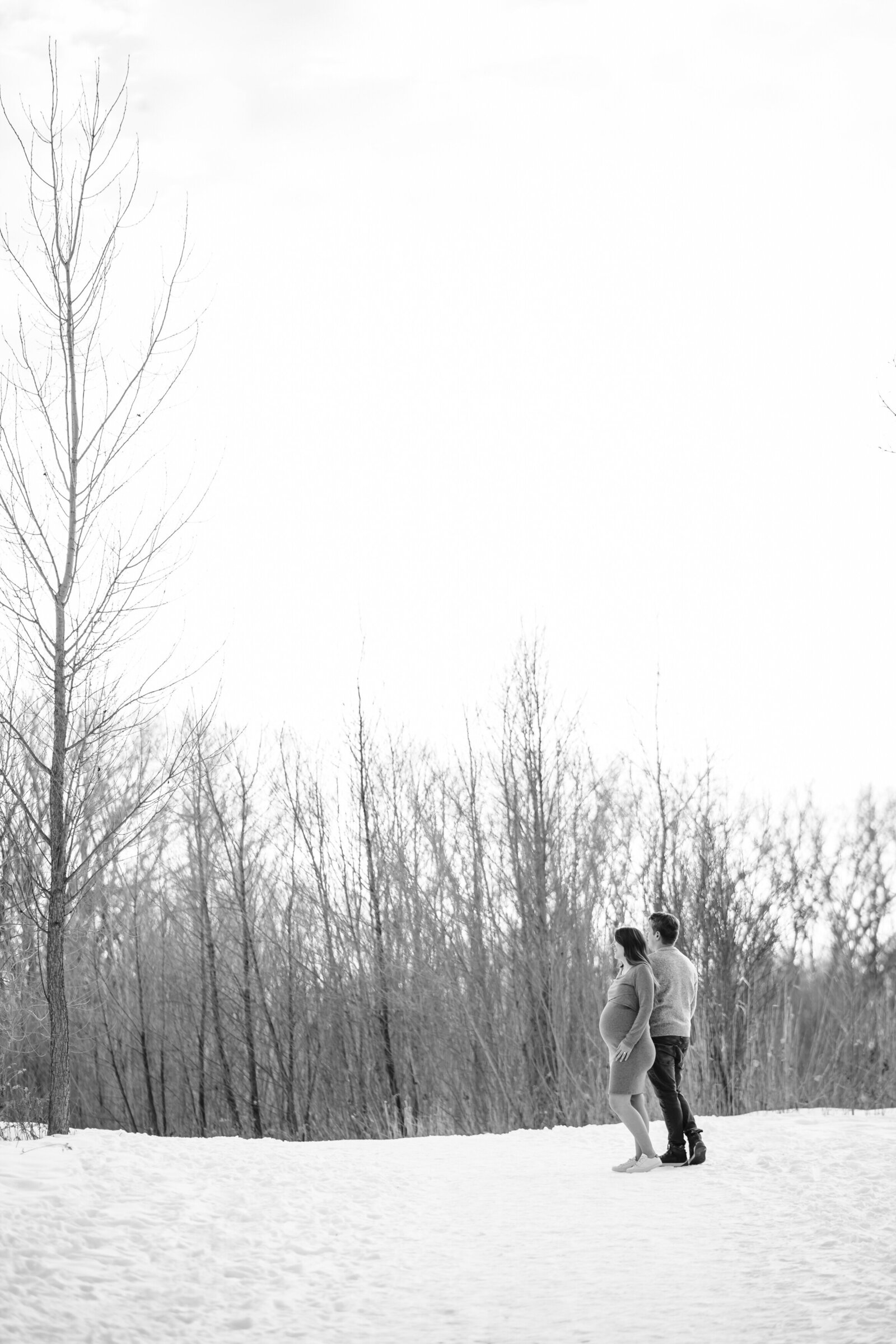 A black and white image of a pregnant woman walking in the snow with her partner. The couple is holding hands, and the barren trees in the background give the scene a quiet, wintery feel.