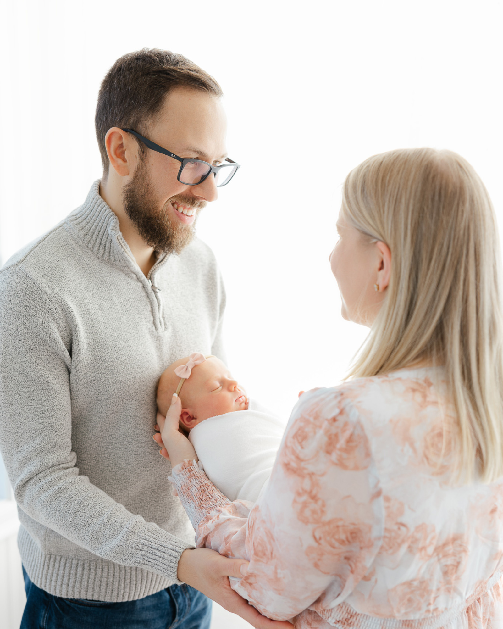 mom in pink floral dress looks at husband smiling and holds newborn baby