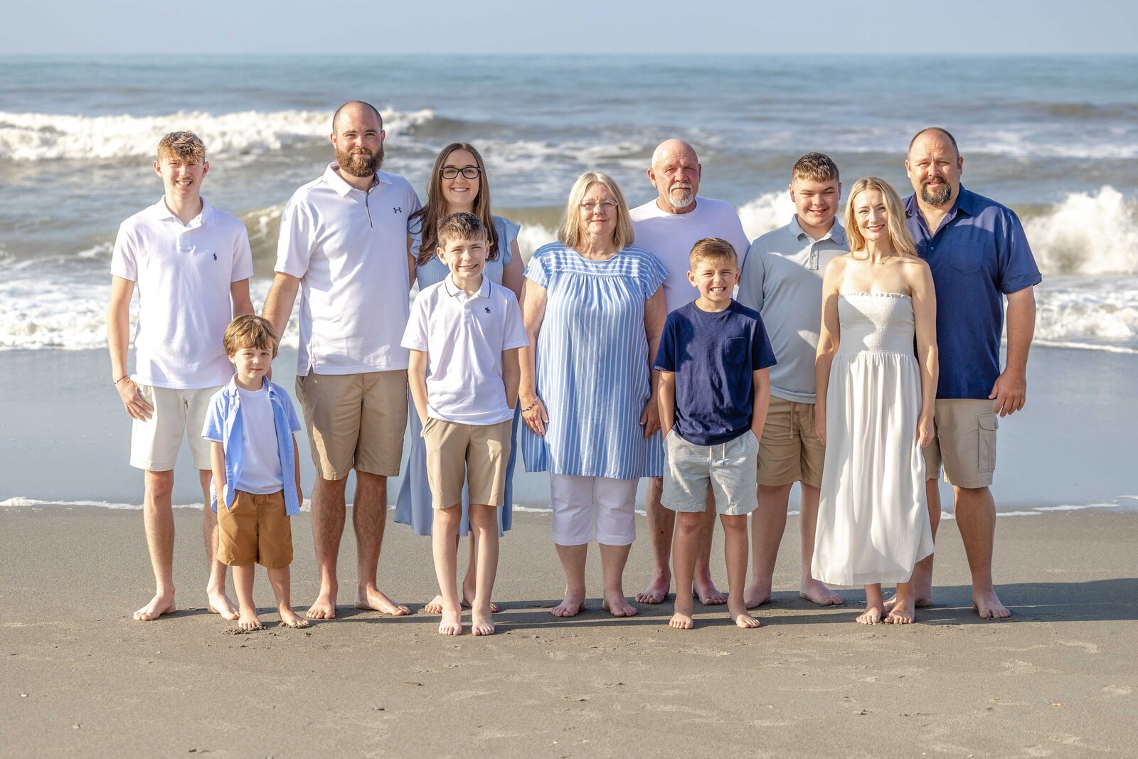 A family poses barefoot on the beach, dressed in blue and white attire. The ocean waves provide a stunning backdrop, capturing the essence of a relaxed family gathering, perfect for artistic family photography in Wilmington, NC.