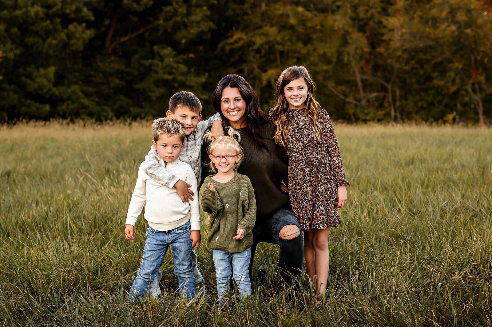 A woman kneels in a grassy field surrounded by four children, their faces bright with smiles. Two boys in light tops sit to the left, while a girl in green stands close by and another in a floral dress graces the right, captured beautifully by a Bloomington family photographer against a backdrop of trees.