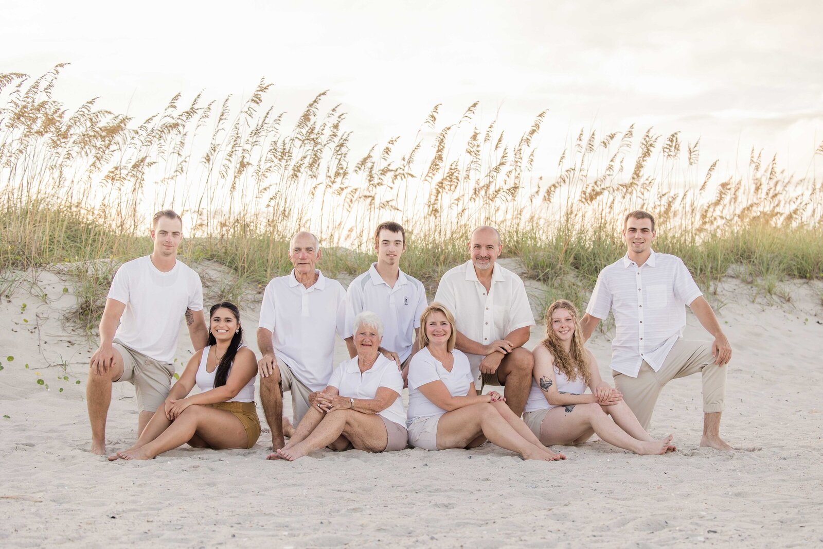 A family sits together on sandy dunes, dressed in elegant white attire with sea oats in the background. The serene setting highlights their close bond, ideal for destination family photographer NC, showcasing timeless and intimate beach portraits.