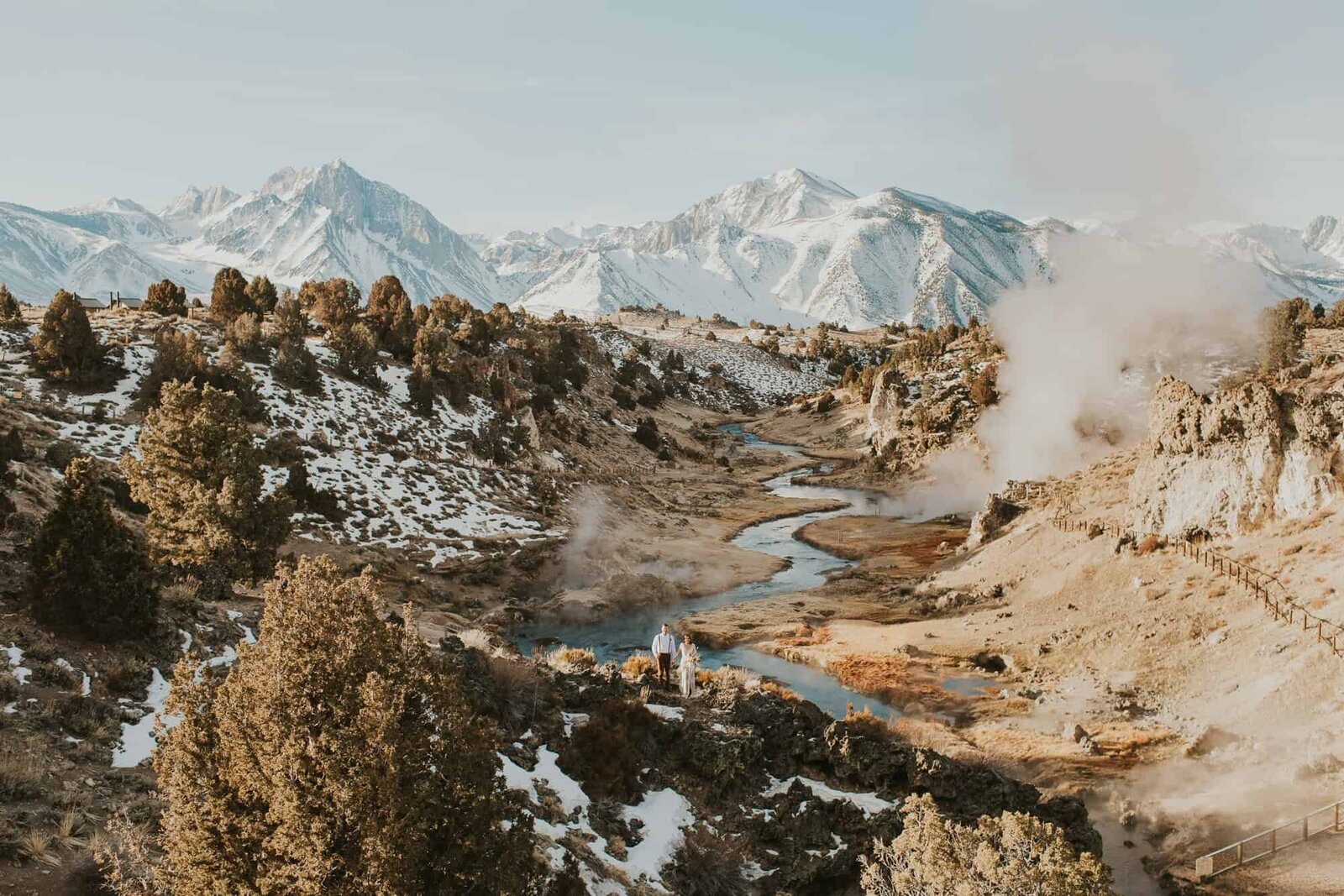 A bride and groom stand above hot creek geological site near Mammoth Lakes, California, with a big view of snow covered eastern sierra mountains behind them. 