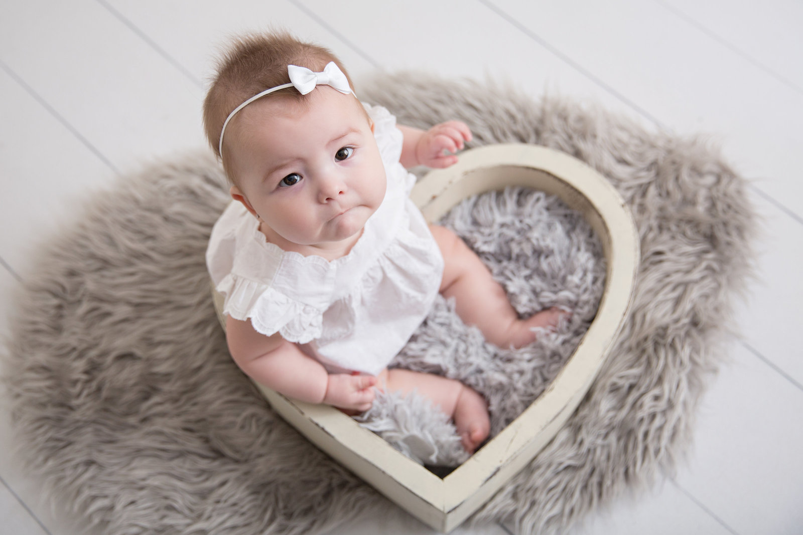 baby girl sitting in white heart bowl with white outfit and bow