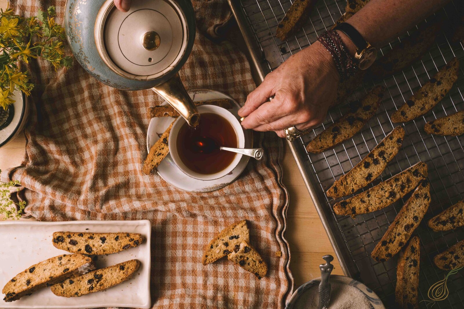 A person gracefully pours steaming tea from a teapot into a delicate cup, capturing a moment of tranquility as biscottis lay on a baking rack.