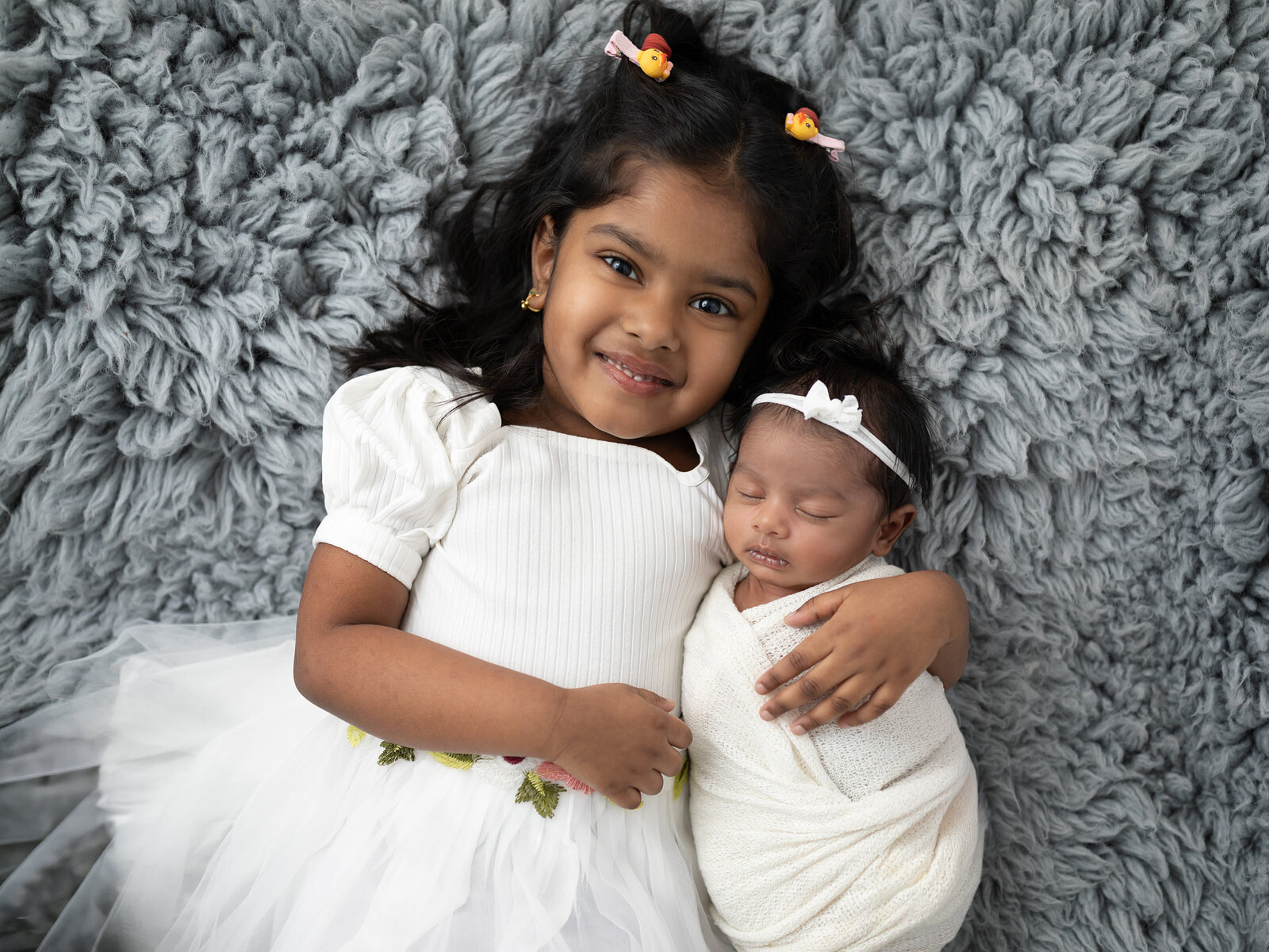 sister laying on gray fur holding newborn baby girl for studio portraits 