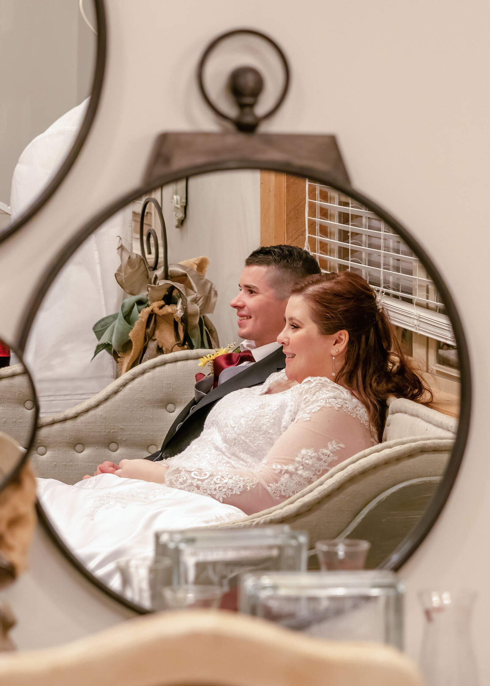 A candid reflection of the bride and groom relaxing together on a vintage couch, captured through a rustic mirror in the Duplin Winery wedding venue bridal suite in Wallace, NC, showcasing authentic moment and elegant decor.