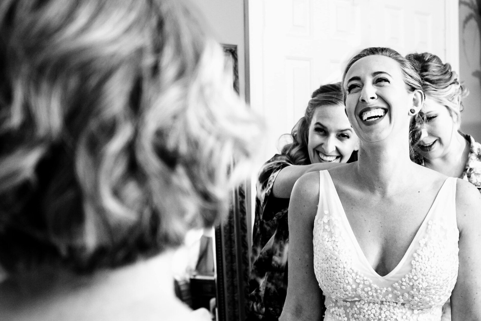 bride laughs as her bridesmaids button up her dress; the mother of the bride looks on in the foreground