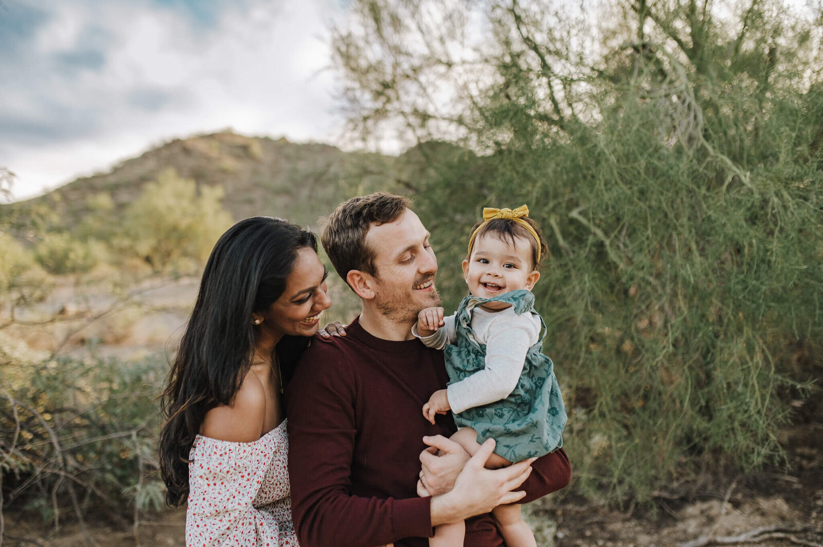 toddler girl smiles at the camera while dad holds her and mom snuggles him from  behind with a mountain desert backdrop