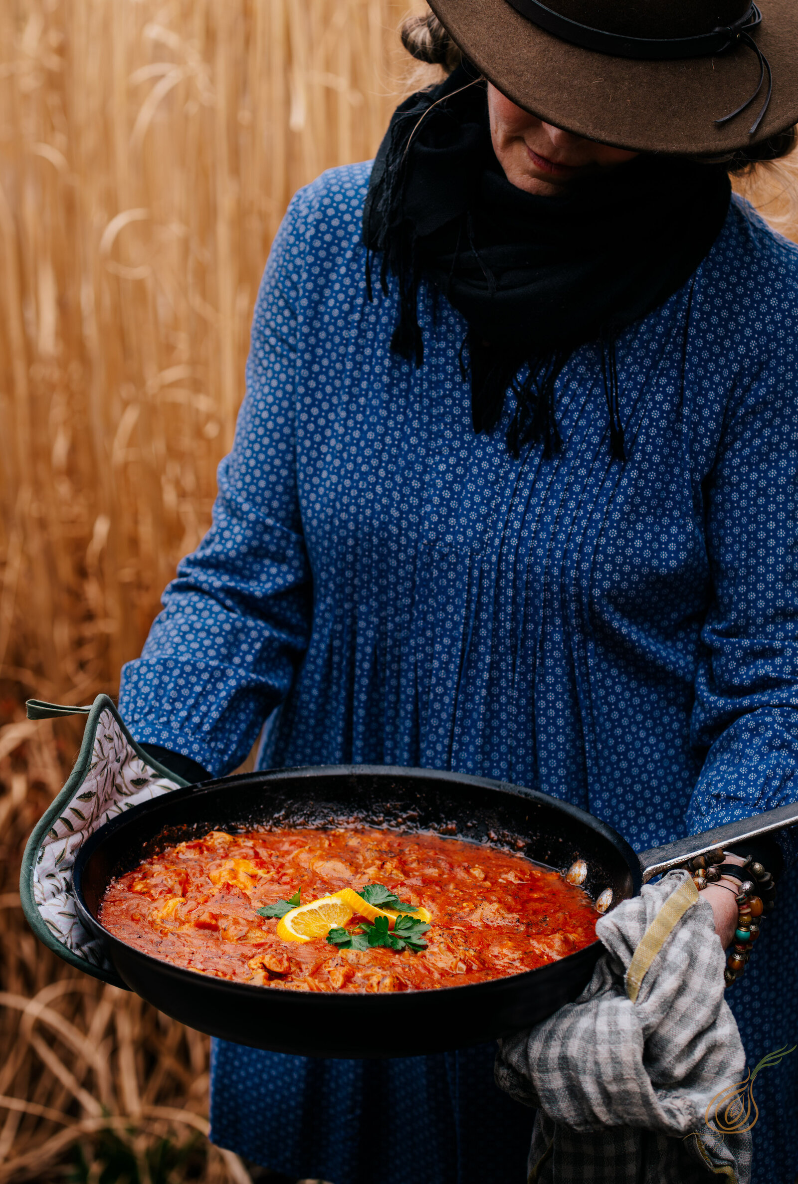A woman in a blue shirt and hat holds a pan filled with tomato sauce, showcasing her cooking skills behind some golden wheat stalks. 