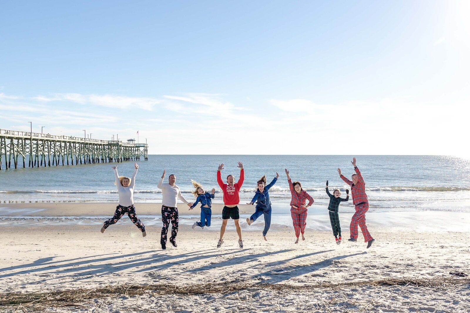 A family joyfully jumps on the beach, dressed in festive pajamas. The bright ocean and pier in the background add to the playful scene, perfect for artistic family photography in Wilmington, NC, capturing fun and energetic holiday moments.