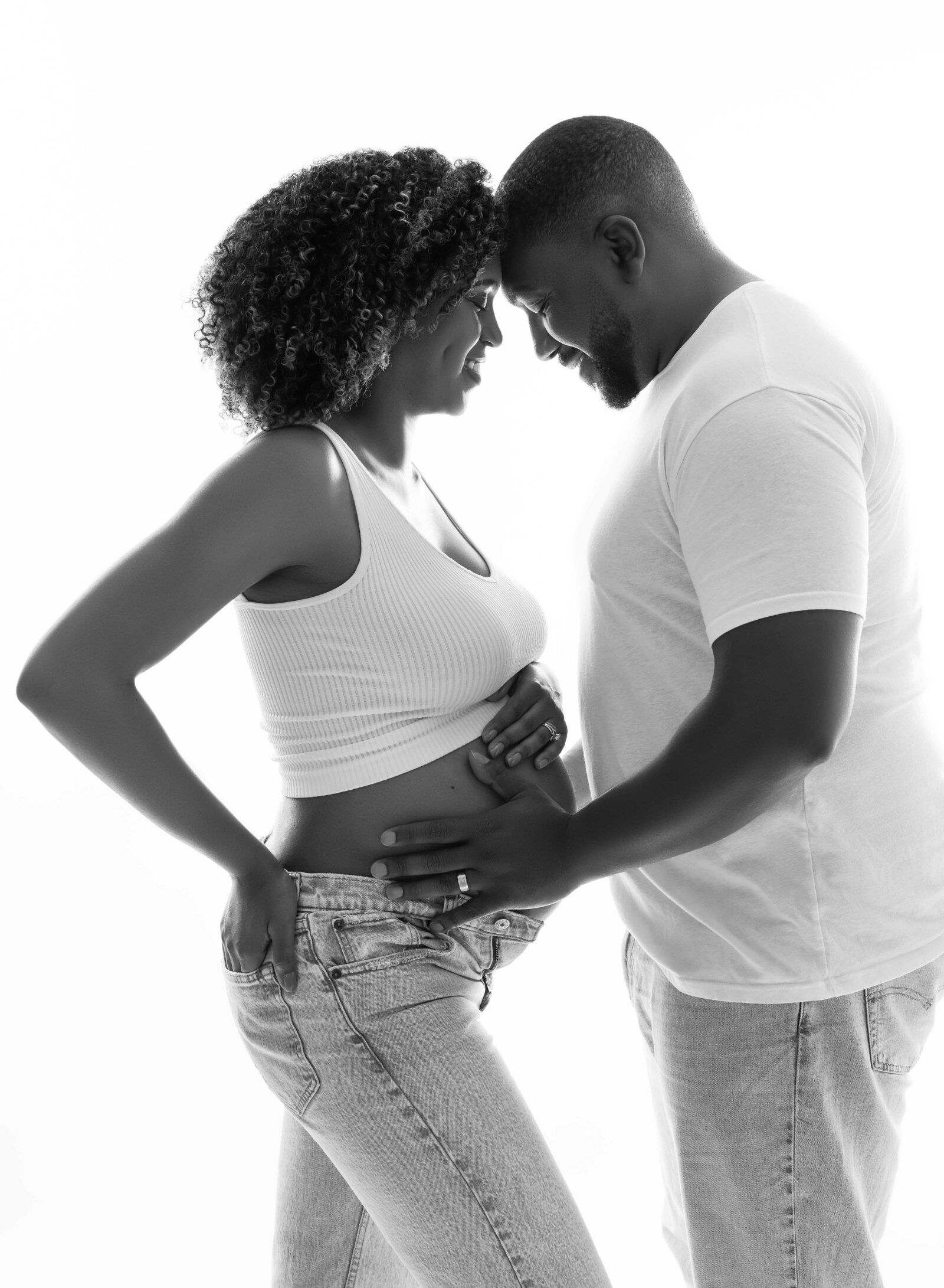 Couple in white shirts and jeans smiling and looking at baby bump. Photographed at Maternity Photo studio in Atlanta.