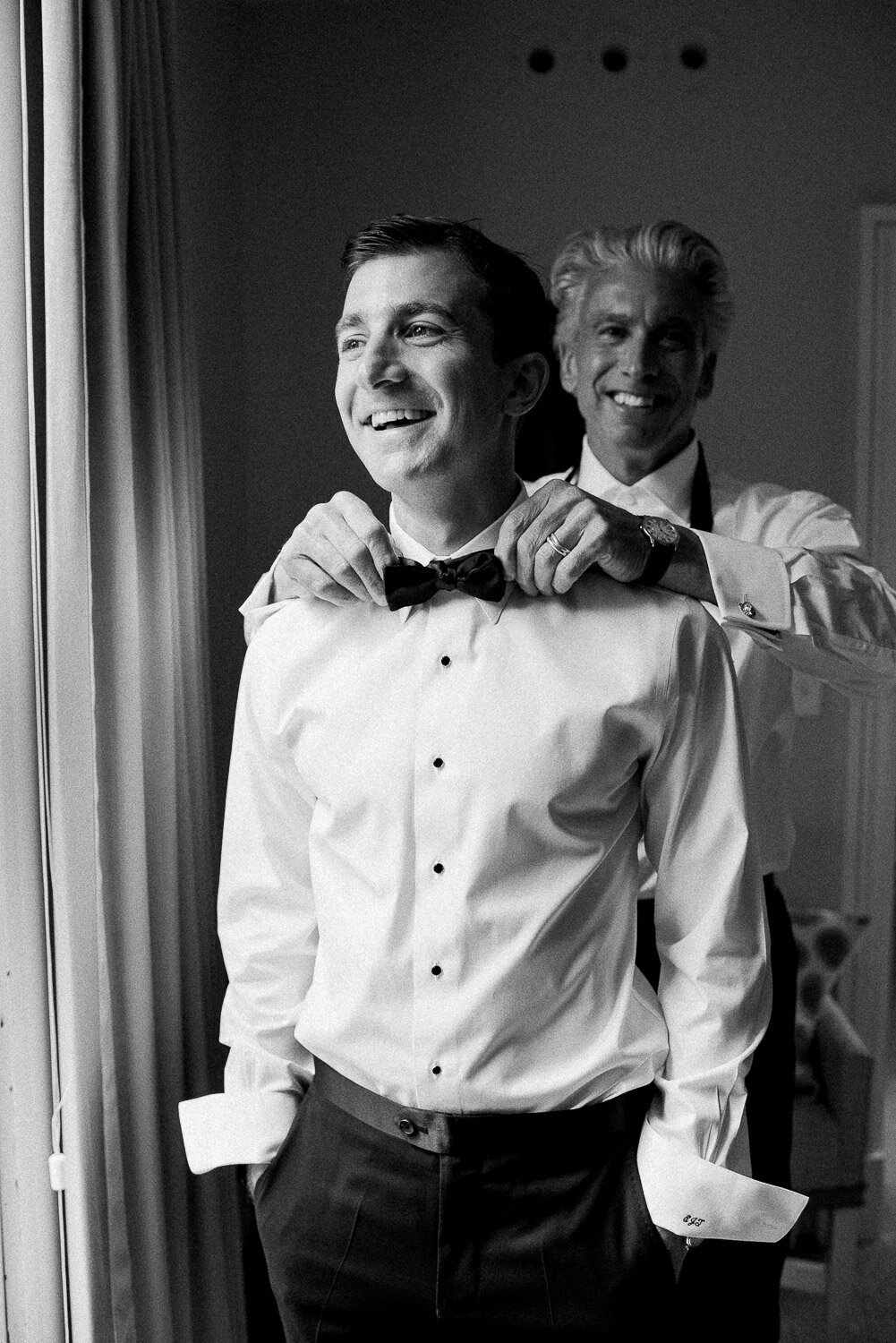 father helping groom with tie in window getting ready