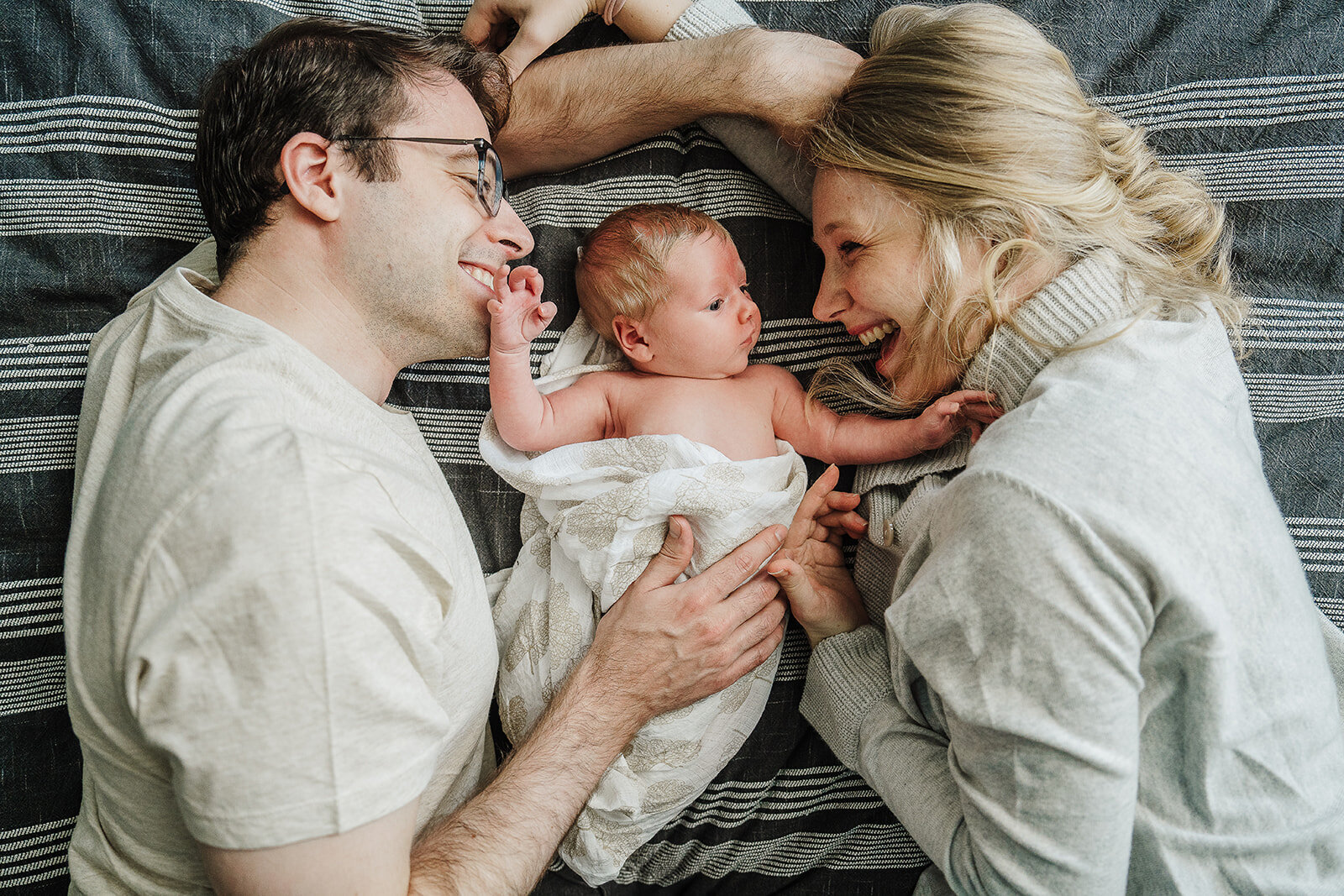 parents laugh looking at newborn while laying on bed