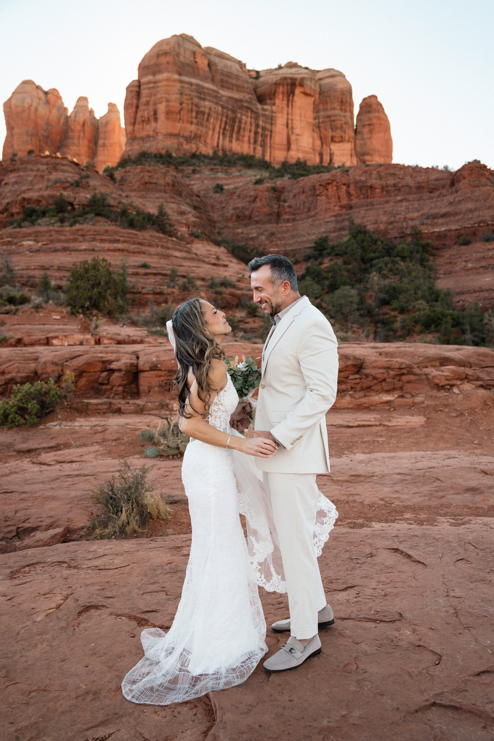 Bride and groom smiling at each other during their elopement at Cathedral Rock in Sedona, Arizona, standing on red rock terrain with dramatic sandstone cliffs in the background.