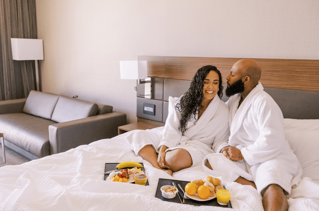 A Black woman with long hair and a Black man sit on a bed in a hotel room. Trays with breakfast food and orange juice are on the bed in front of them.