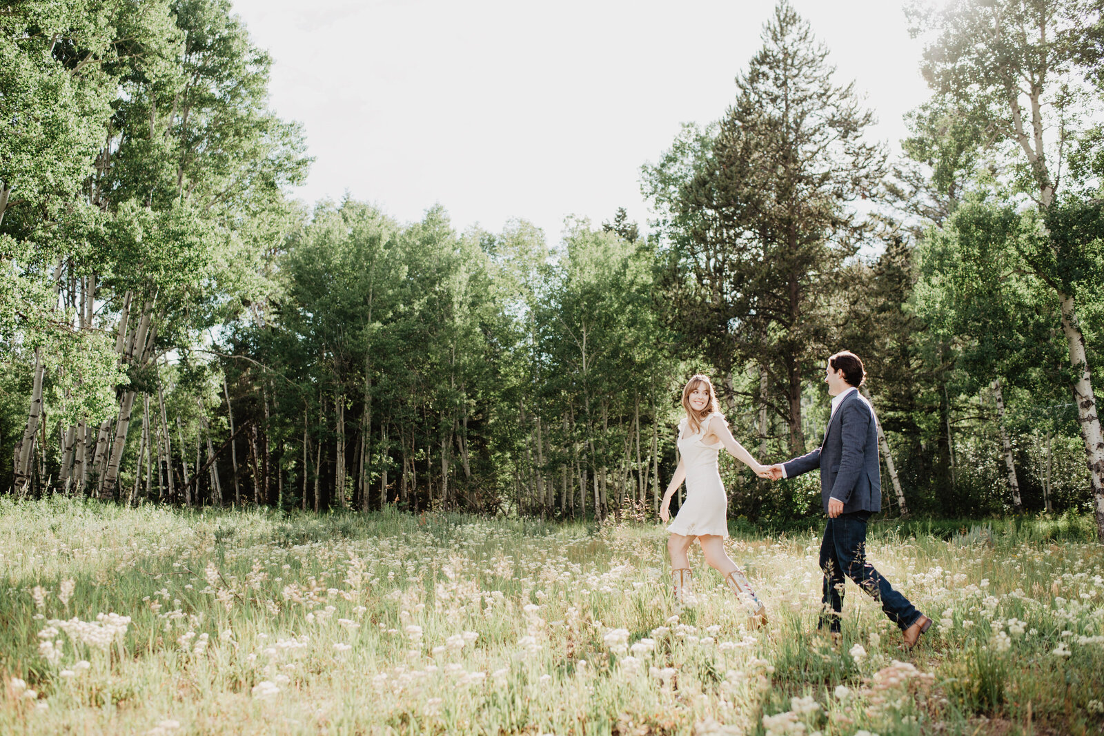 couple holding hands and walking together through a wildflower field in a meadow with a forest surrounding the field as the woman looks back to her fiance and smiles to him