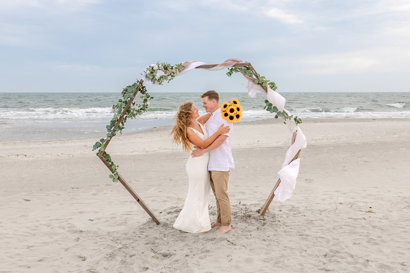 The bride and groom embrace under a circular beach ceremony arbor adorned with greenery and white fabric, captured during a romantic Ocean Isle Beach, NC wedding, blending coastal elegance and natural beauty.