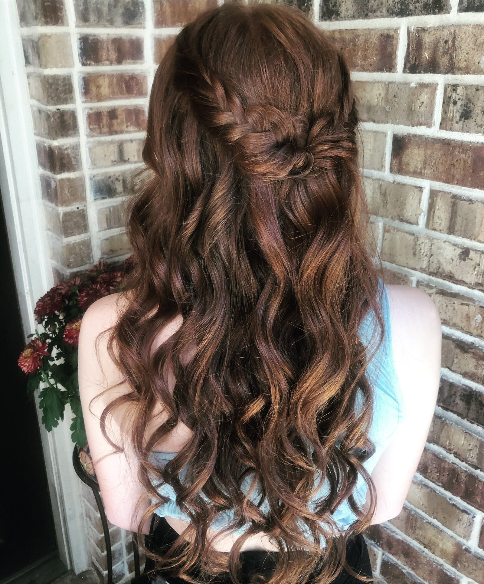 Person with long, wavy auburn hair styled in a braided half-updo against a brick wall background, capturing the elegance of a bridal beauty experience Texas can offer.