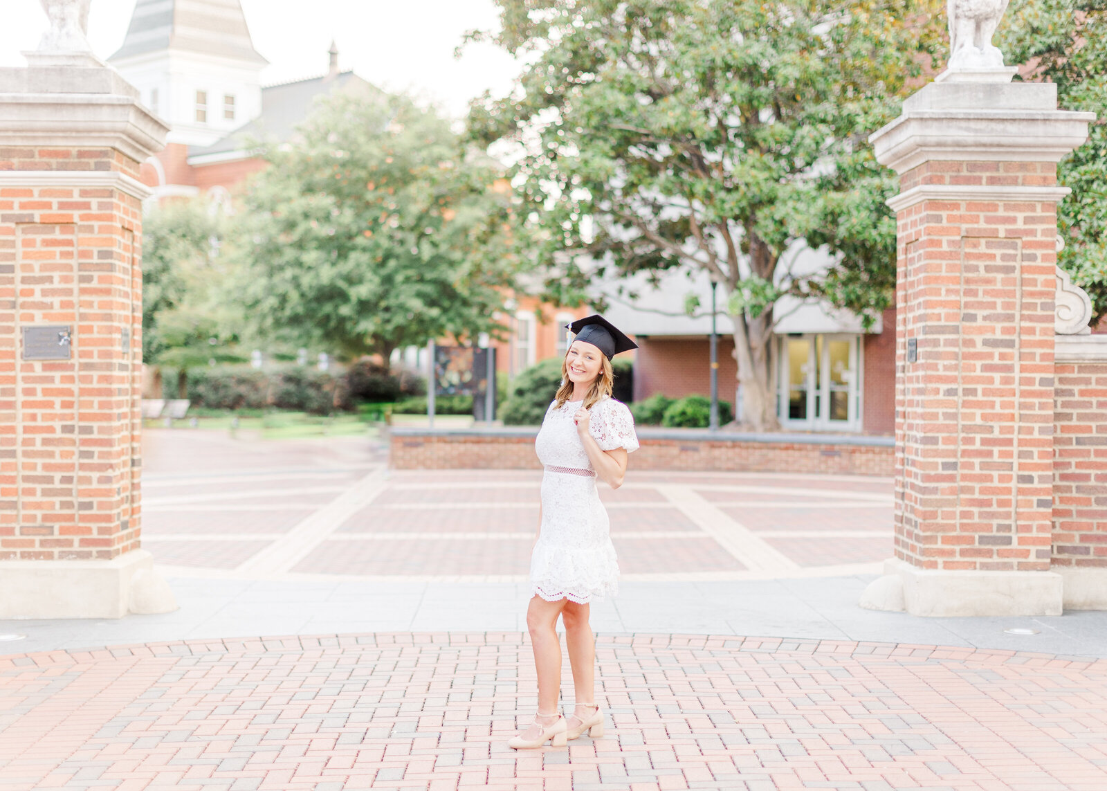 Shelby smiling and posing at a university campus gate, wearing her graduation cap and white dress, photographed during her senior portraits.