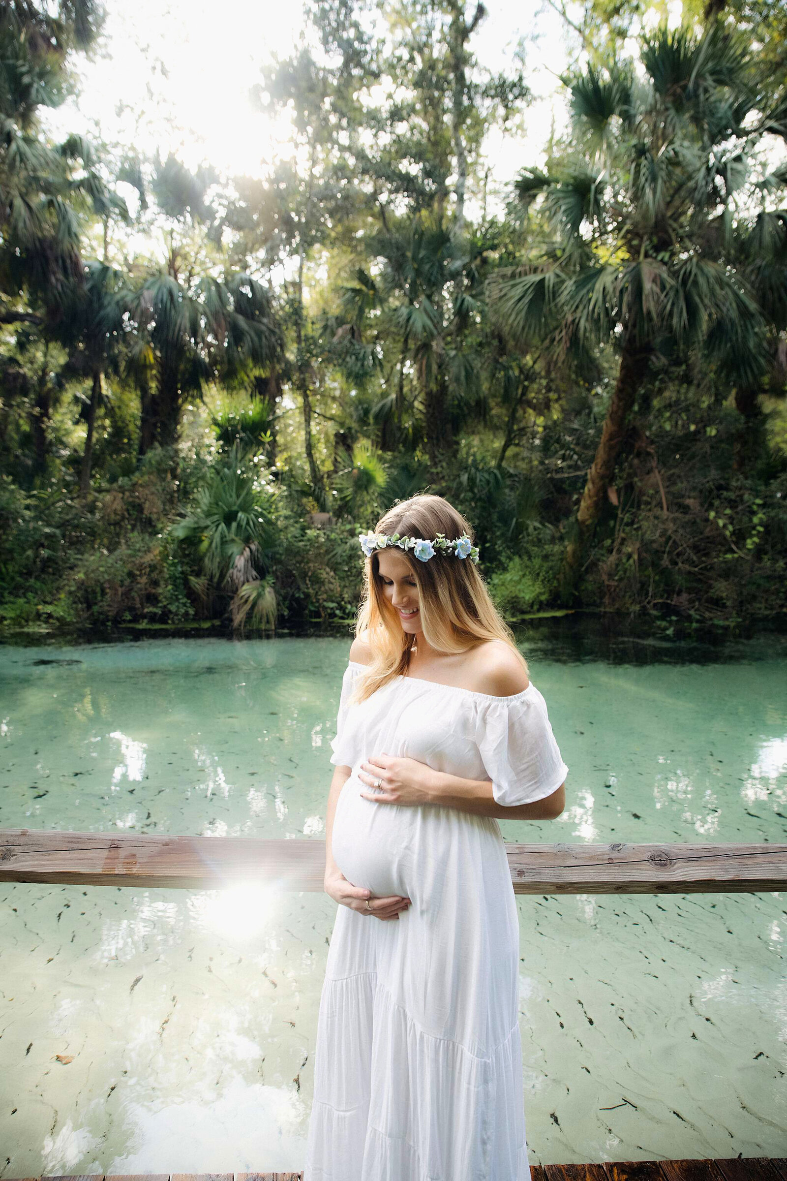 A pregnant woman in a flowing white dress poses on a wooden bridge with water in the background, photo by Bia Schaefer.