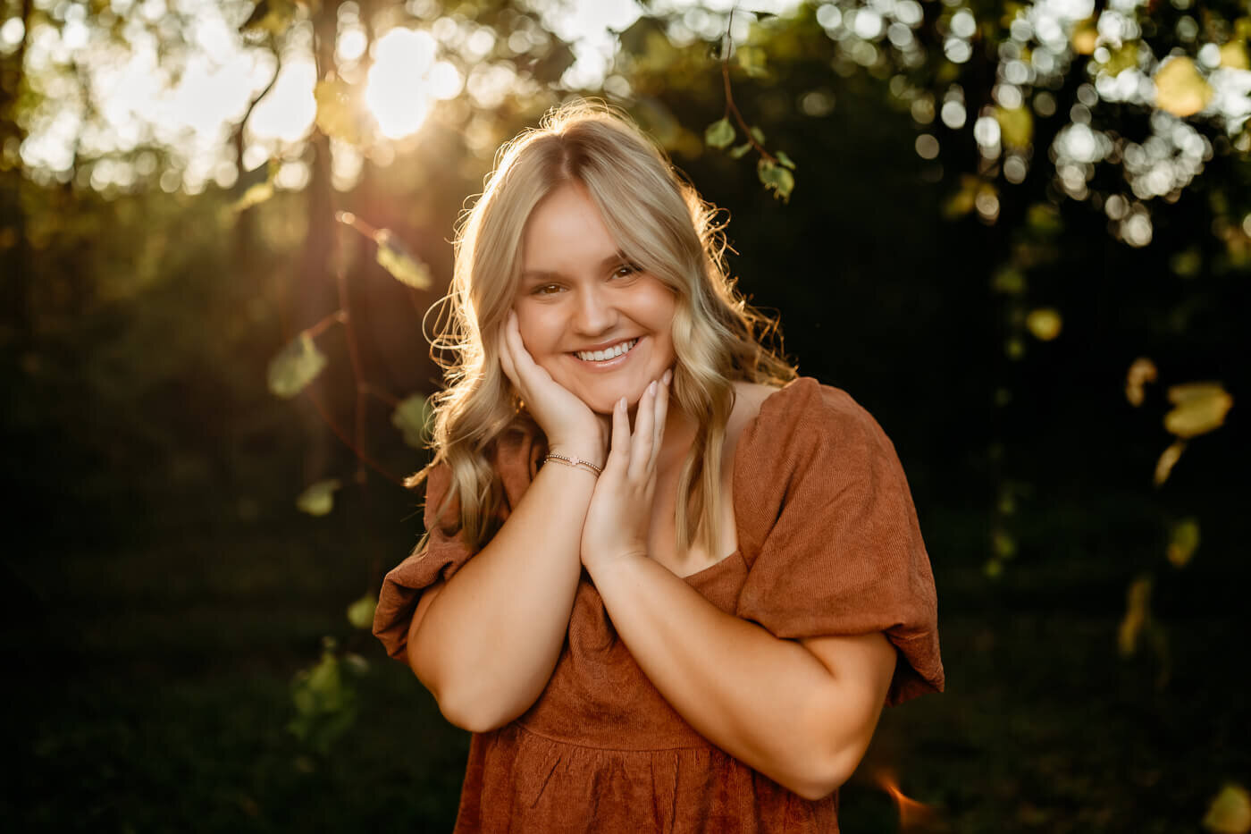 A person with long hair and a brown dress smiles while standing outdoors. The sun shines brightly through the trees in the background, creating a warm and serene atmosphere.
