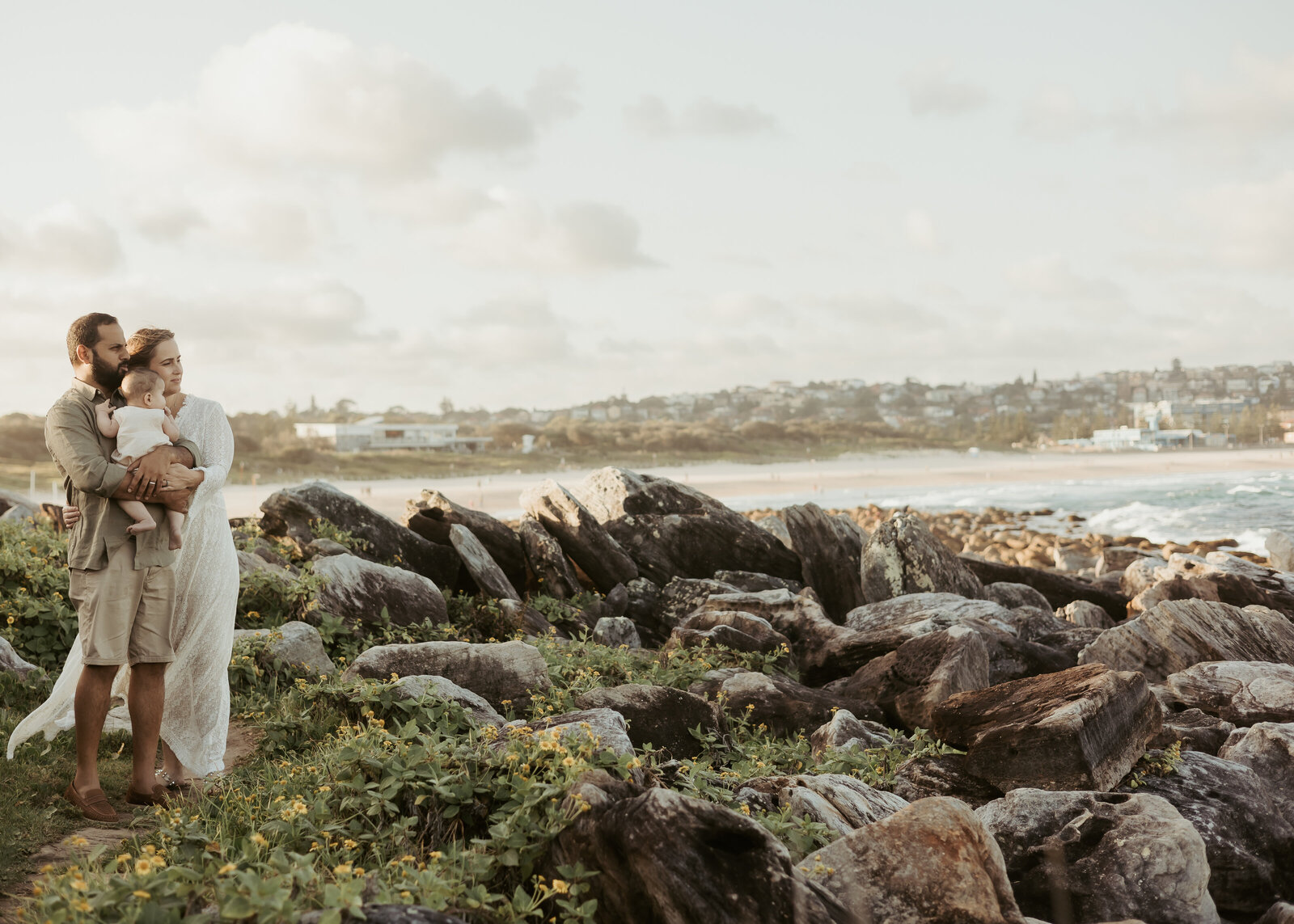 A six month milestone photography family session with baby and parents posing together at rocks near the beach.